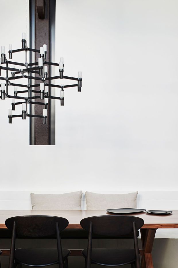 A refined dining room featuring a long dark timber dining table with black sculptural chairs, neutral upholstered cushions, and a dramatic black multi-arm pendant light mounted in front of a dark vertical wall panel. Minimal white walls and restrained tableware create a sophisticated, high-contrast composition.
