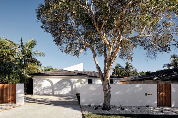 Street-facing view of a low-rise contemporary residence with a white rendered facade, tiled hipped roof, arched recessed detailing, timber slat gates, and a broad pale gravel forecourt. A mature tree and tropical planting occupy the frontage, with strong sunlight and clear blue sky.