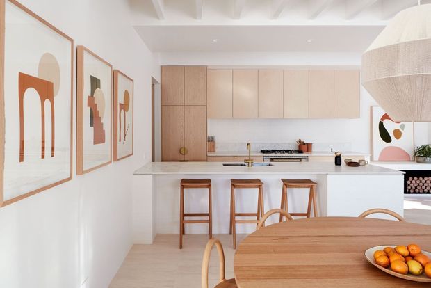 Bright open-plan kitchen and dining area with a large white island, pale timber cabinetry, integrated cooking zone, timber bar stools, and a rounded timber dining table. The room features white walls and ceiling beams, framed abstract artwork, light timber flooring, and soft contemporary styling with warm natural accents.