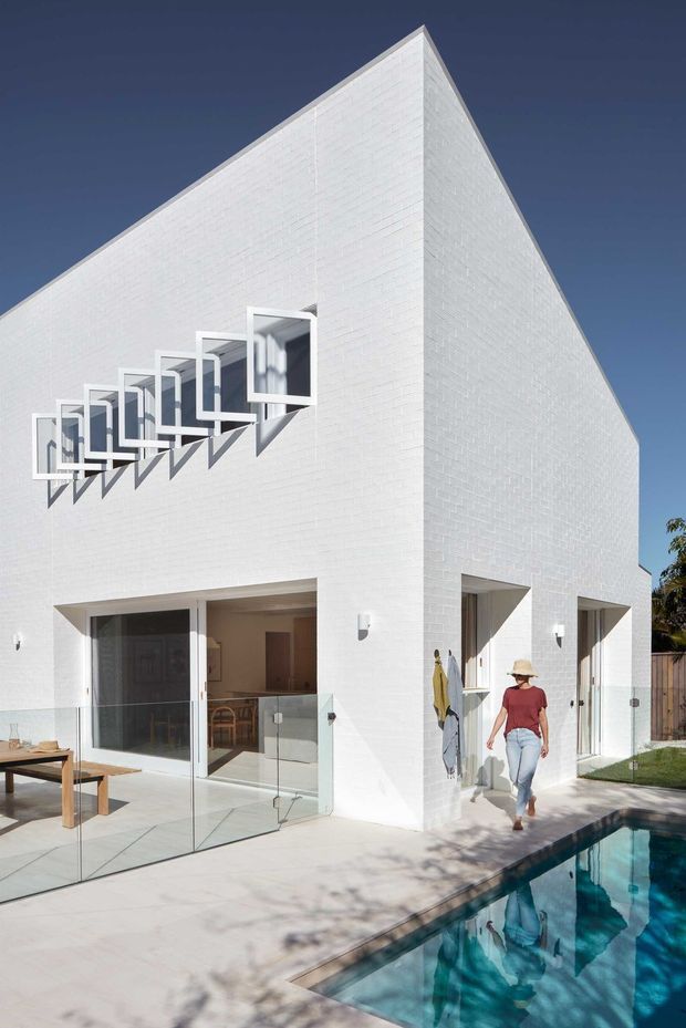 A bright contemporary white-brick residence viewed from the pool terrace. The sharply angular, wedge-like building has a largely blank side facade, a row of projecting square window boxes with deep reveals, and large glazed openings at ground level. A pale paved terrace with outdoor dining furniture borders a rectangular swimming pool, while a small portion of the interior is visible through the glazing.