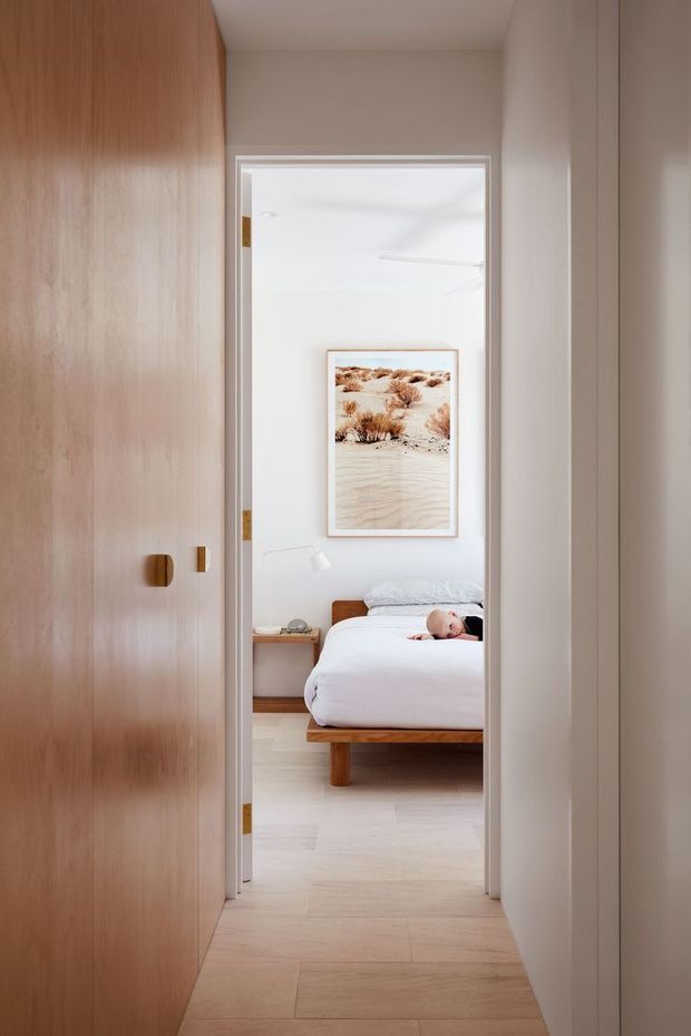 View through a hallway doorway into a bright, softly styled bedroom. The bedroom contains a low timber bed with white bedding, a small bedside table and lamp, and a framed desert landscape print. A full-height warm timber built-in wardrobe or wall of cabinetry lines the left side of the foreground, while pale timber flooring and warm off-white walls create a calm, minimal interior.