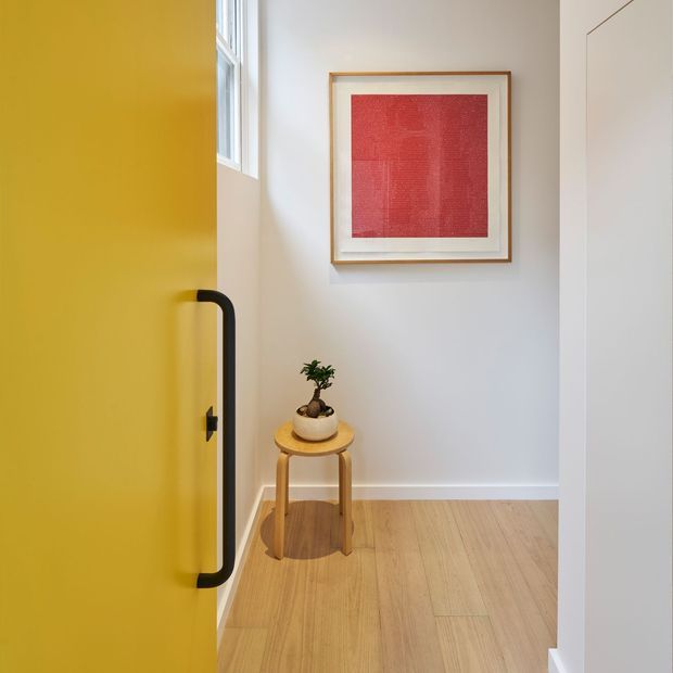 A bright, minimalist entryway viewed through a partially open mustard-yellow door with a black vertical pull handle. The space has pale walls, light timber flooring, a small round timber stool with a potted plant, a high narrow window, and a framed red artwork. Clean architectural lines and warm natural light create a refined, welcoming feel.