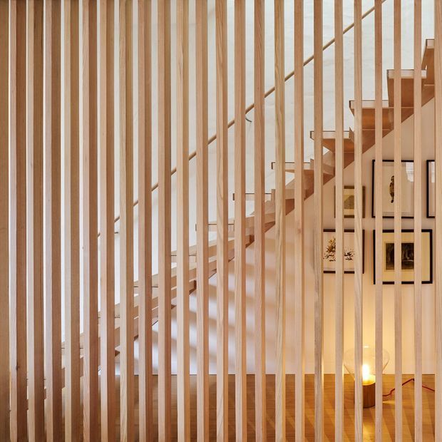 Interior view of a light-filled stairway screened by closely spaced vertical timber battens. A pale timber staircase rises diagonally behind the slatted divider, with timber flooring, white walls, framed artwork, and warm accent lighting visible beyond.
