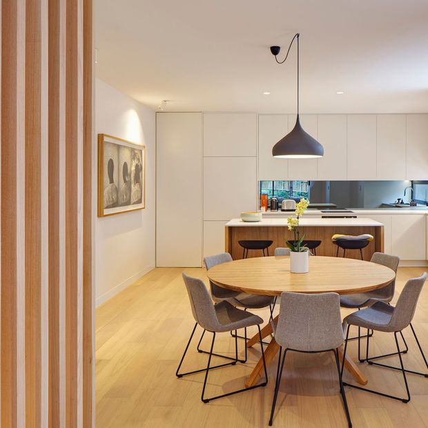 A warm, contemporary open-plan dining and kitchen interior. A round timber dining table with upholstered chairs occupies the foreground, while a streamlined white kitchen with integrated cabinetry, island seating, and a reflective backsplash sits behind it. Light timber flooring, white walls and joinery, vertical timber wall detailing, framed artwork, and a large dark sculptural pendant create a refined, inviting space.