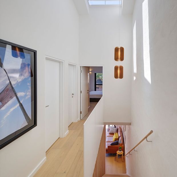 Bright, narrow upper-level hallway and stair landing with white walls, pale timber flooring, a high skylight, tall side windows, two suspended timber pendant lights, and a stair descending to a living area below. A bedroom is visible through the open doorway at the end of the hall, while framed artwork lines the left wall.