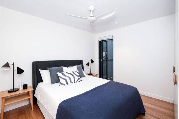 Bright, contemporary bedroom with a white upholstered or fabric headboard, white bedding, navy-blue throw, and patterned accent cushions. Light timber flooring and matching bedside tables add warmth, while black wall-mounted and table lamps provide contrast. A ceiling fan, built-in wardrobe, and glazed doorway are visible.