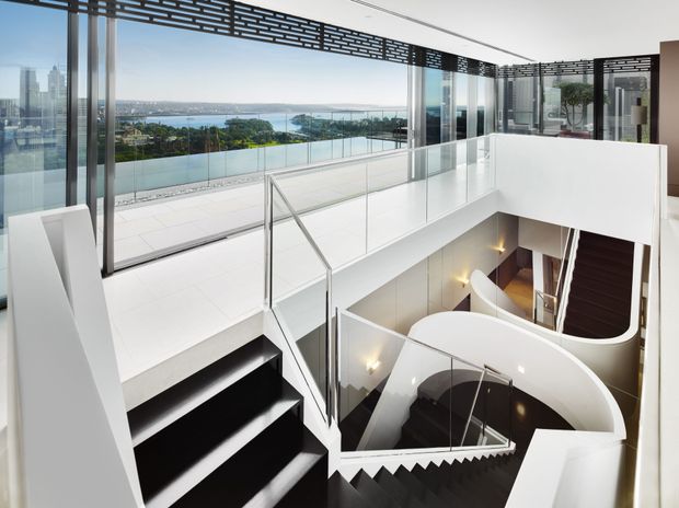 A dramatic contemporary multi-level interior featuring a sculptural staircase with black steps, white curved balustrades, glass railings, and a double-height void. Floor-to-ceiling glazing opens to expansive waterfront and city views, while a perforated dark overhead screen adds architectural detail.