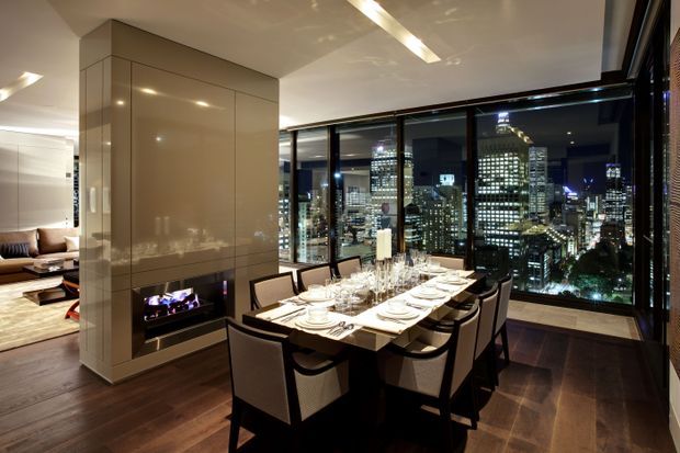 A sophisticated high-rise apartment dining area at night, featuring a formal dining table set for eight beside floor-to-ceiling glazing with illuminated city views. A glossy beige tiled fireplace volume separates the dining space from a partially visible living room, while dark timber flooring, black-framed glazing, and linear ceiling lighting create a refined urban atmosphere.