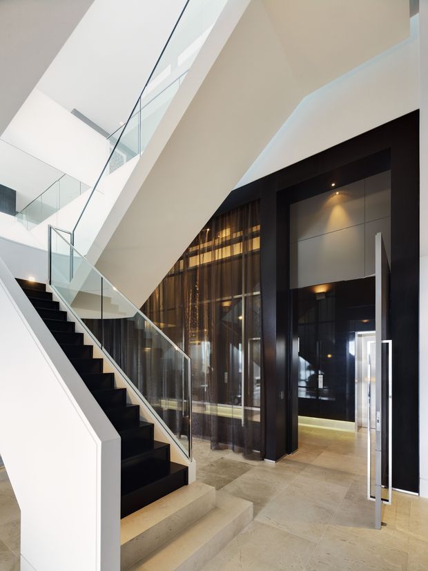 A dramatic contemporary interior entry hall featuring a sculptural black-and-white staircase, glass balustrades, polished light stone flooring, and a double-height void. Dark framed glazing and sheer curtains line the adjacent space, while a tall black-framed entrance with long metallic handles creates a strong architectural focal point. The space is brightly lit with recessed and concealed lighting.