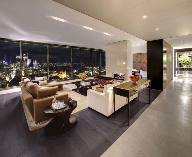 A sophisticated high-rise living room at night with expansive floor-to-ceiling glazing overlooking city lights. The room features a large neutral sectional seating arrangement, sculptural dark coffee tables, a light upholstered armchair, a slim console table, dark timber flooring, pale stone or tile flooring in the circulation area, recessed ceiling lighting, and dark built-in cabinetry. A hallway and greenery are visible beyond the living area.