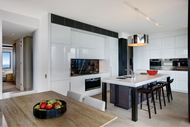 A bright, contemporary open-plan kitchen and dining area with glossy white floor-to-ceiling cabinetry, integrated black appliances, a central island with an undermount sink and seating, and a timber dining table in the foreground. A dark textured splashback and linear pendant lighting add contrast to the otherwise crisp white interior.