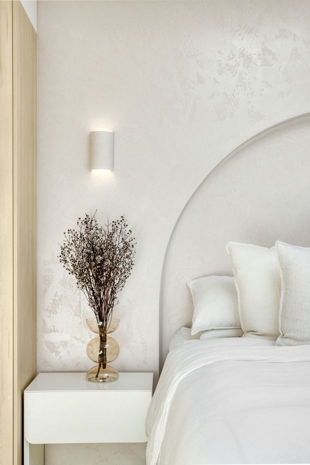 A close-up of a serene, softly styled bedroom corner. A white bed with layered pillows sits beside a minimalist wall-mounted white bedside cabinet, topped with a translucent glass vase holding dried botanical stems. The room features a warm light-wood vertical panel, textured off-white plaster walls, a curved recessed architectural bedhead detail, and a simple wall sconce. The palette is predominantly warm white, cream, and pale timber, creating a calm contemporary atmosphere.