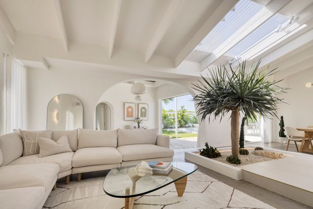 Bright, airy open-plan living room with a large cream sectional sofa, glass coffee table, arched wall openings, white exposed ceiling beams, and a dramatic indoor planter containing tall sculptural palms. Skylights and large windows bring in abundant daylight, with a dining area visible in the background.