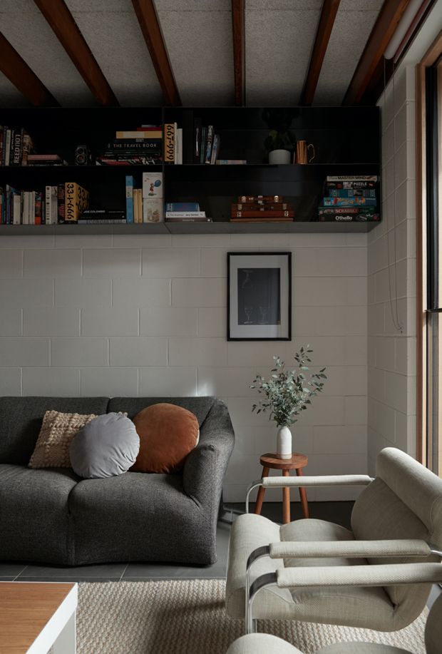 A cozy living room with a dark charcoal sofa, textured cushions, a cream upholstered lounge chair, and a small timber side table with a vase. The room features a pale painted concrete-block wall, a black built-in open bookshelf filled with books and games, exposed dark timber ceiling beams, framed wall art, and large vertical glazing at the right. Natural light creates a warm, relaxed atmosphere.