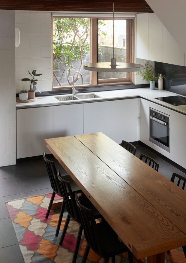 A bright, compact contemporary kitchen-dining space with white handleless cabinetry, pale benchtops, integrated stainless-steel appliances, a dark tiled floor, and a large natural timber dining table surrounded by black chairs. A timber-framed window brings in daylight above the sink, while a dark timber slatted ceiling and colourful patterned rug add warmth and contrast.
