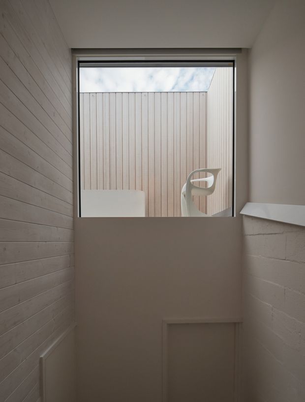 A narrow, softly lit stairwell or landing with pale painted horizontal timber wall panelling on the left, a white-painted masonry wall on the right, and a large black-framed window at the upper end. The window looks onto a close vertical timber privacy screen and reveals a sculptural white outdoor chair or object beyond. The space has a restrained, minimalist architectural character with neutral finishes and a partially visible stair or lower-level opening.