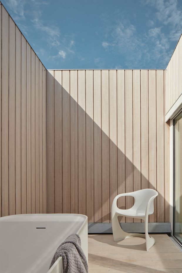 A bright, minimalist outdoor bathing terrace enclosed by tall vertical timber-clad privacy walls. The image shows the corner of a freestanding white bathtub, a sculptural white chair, pale timber decking, and a large glass door. Strong sunlight creates a dramatic diagonal shadow across the warm wood panelling, with open blue sky above.