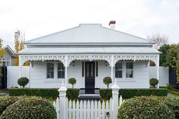 Front-on exterior view of a single-storey heritage-style home with pale horizontal weatherboard cladding, a broad hipped corrugated metal roof, ornate white lacework verandah detailing, dark timber entry door, symmetrical windows, and a manicured front garden enclosed by a white picket fence.