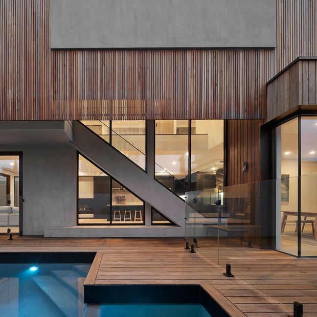 Contemporary multi-level residential courtyard viewed from a timber deck beside a swimming pool. The building combines dark vertical timber cladding, raw concrete walls, extensive black-framed glazing, and a prominent external stair with a strong diagonal profile. Warm interior lighting and visible living spaces create a refined indoor–outdoor connection.