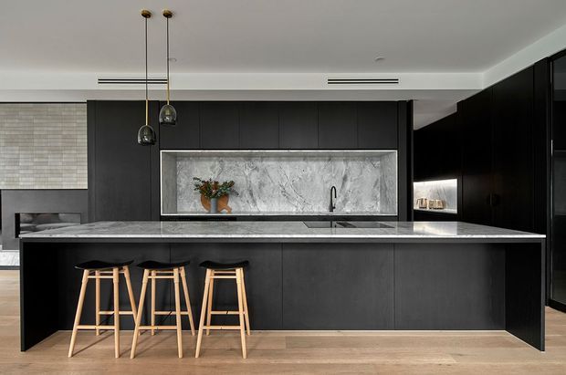 A contemporary kitchen dominated by a large black island with a pale stone benchtop and three timber bar stools. Full-height dark cabinetry frames a dramatic grey-and-white marble-look splashback, with integrated appliances, pendant lights, light timber flooring, and a partial view toward an adjacent living area.