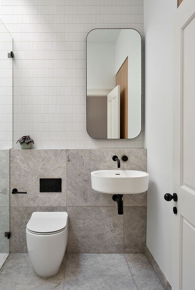 Compact contemporary bathroom featuring a wall-hung white toilet, floating white basin, matte black tapware and flush plate, grey stone-look wall and floor tiles, and a vertically ribbed white tile feature wall. A rounded-rectangle black-framed mirror reflects a paneled interior door. The space has a clean, refined, neutral design with strong black accents.