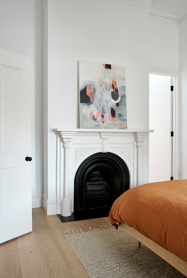 Bright, refined bedroom featuring a decorative white carved fireplace mantel with a black cast-iron insert, white paneled doors, ornate ceiling cornices, pale timber flooring, and a rust-orange upholstered bed. A soft abstract artwork hangs above the fireplace, with a neutral woven rug grounding the room.