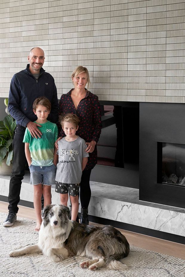 A family and dog pose in a contemporary living room beside a large black built-in fireplace. The room features a full-height horizontally tiled or panelled feature wall, a marble-look hearth and dark timber flooring, with a neutral textured rug and indoor plant.