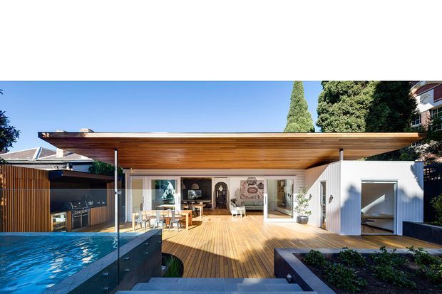 Contemporary single-storey rear extension opening onto a timber deck and swimming pool. The pavilion has a broad flat overhanging roof with exposed timber soffit, extensive glazed sliding doors, white vertical cladding, and an integrated outdoor kitchen at left. The image is photographed from the pool area, with the deck and landscaped garden framing the indoor-outdoor living space.