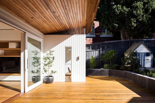A sunlit timber-clad outdoor deck and covered terrace adjoining a contemporary interior. The space features warm timber decking and a timber-lined ceiling, white vertical wall cladding, large glazed folding or hinged doors, built-in timber shelving visible inside, potted plants, and a curved raised garden bed beside a dark privacy fence. A small garden shed is visible beyond the terrace.
