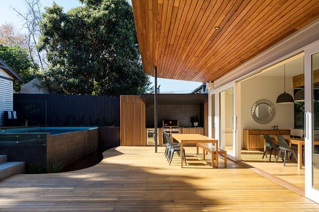 A sunlit contemporary outdoor entertaining area with a broad timber deck and timber-lined covered ceiling. Large glazed sliding and folding doors open into a dining and kitchen area, creating a strong indoor-outdoor connection. The deck includes an outdoor dining setting, built-in barbecue or serving area, dark privacy fencing, and a partially visible rectangular pool or spa at the side.