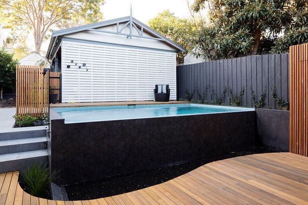 A compact backyard pool area with a raised rectangular swimming pool clad in a dark geometric feature surface. The pool sits beside a warm timber deck and short stairway, with dark vertical fencing, timber screening, planted borders, and a small gabled outbuilding with a white roller door in the background. The setting is landscaped, private, and contemporary.