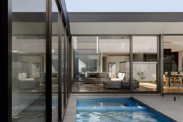 Contemporary courtyard interior with a compact swimming pool surrounded by dark grey tiled paving. Extensive black-framed floor-to-ceiling glazing reflects and reveals a bright living area, dining space, and double-height or high-ceilinged volumes. The composition emphasizes clean rectilinear forms, layered indoor-outdoor living, and a restrained neutral palette.