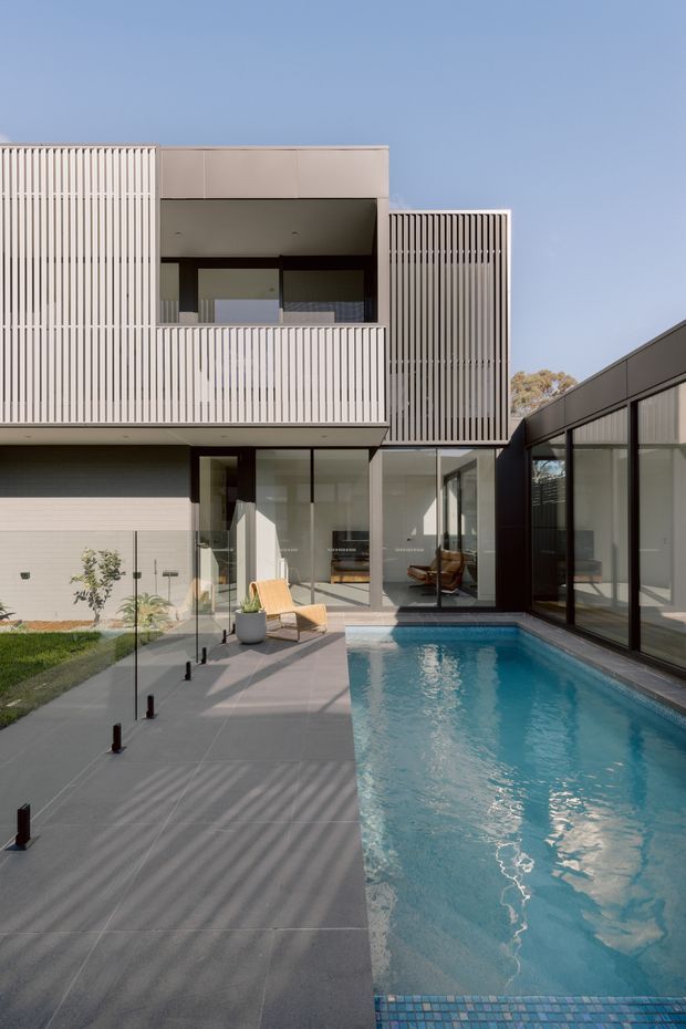 A contemporary multi-level residential house arranged around a narrow rectangular swimming pool and paved courtyard. The facade combines pale vertical slatted screening, smooth light panels, dark window and door frames, and extensive floor-to-ceiling glazing. A covered upper-level balcony opens toward the pool, while the ground floor connects directly to the outdoor area. Strong linear shadows fall across the grey paving in bright daylight.