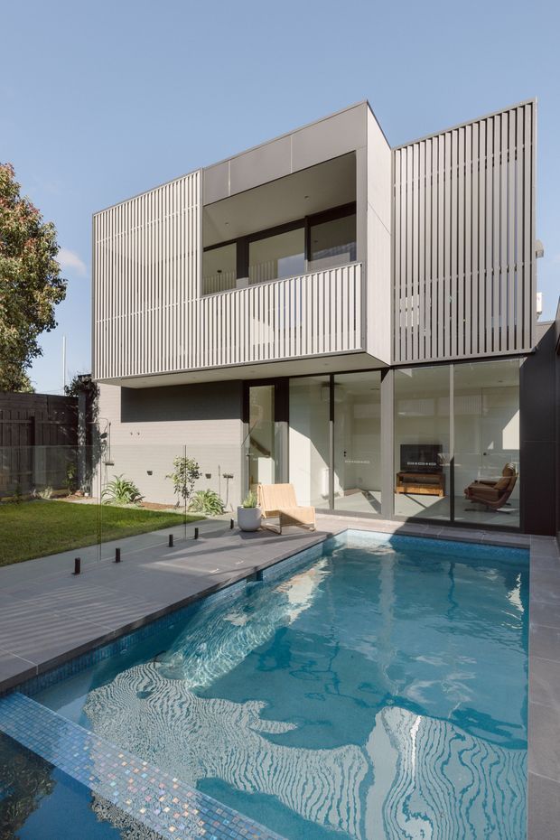 A contemporary multi-storey residence viewed from the backyard, featuring a long swimming pool bordered by a paved deck and lawn. The house has a clean geometric form with pale rendered walls, extensive glazing, and prominent vertical slatted screening on the upper level and side facade. Interior living areas are visible through the ground-floor glass doors.