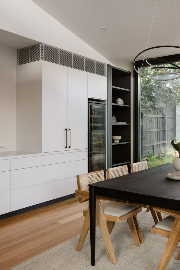 A refined open-plan dining area with a sleek white built-in kitchen wall, integrated wine refrigerator, dark open shelving, and a large black dining table surrounded by woven-back timber chairs. Warm timber flooring, a neutral rug, sculptural pendant lighting, and full-height glazing create a contemporary, softly layered interior.