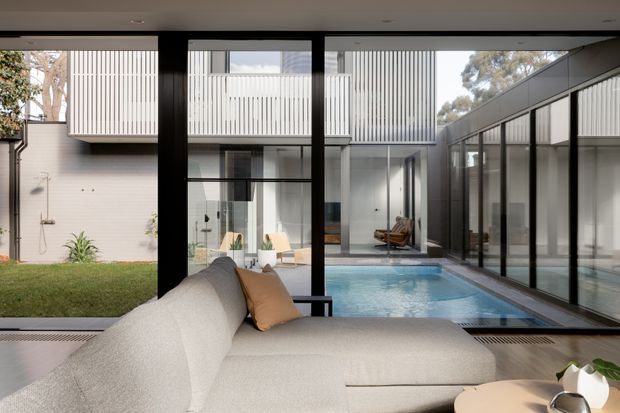 A refined contemporary living room opens through extensive black-framed glazing onto a private courtyard with a narrow swimming pool, lawn, and outdoor shower. The room features a large cream sectional sofa, warm timber flooring, and a low round coffee table. Across the courtyard, white vertically screened upper-level walls and glass-lined circulation spaces create a tranquil, light-filled indoor-outdoor composition.