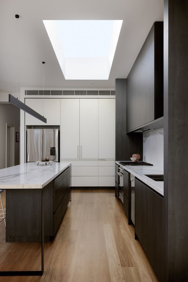 A bright, contemporary galley kitchen with a large central island, dark timber cabinetry, white integrated cabinetry, white veined stone benchtops, stainless-steel appliances, and a large ceiling skylight. Light timber flooring and a linear pendant light create a refined, high-contrast interior.