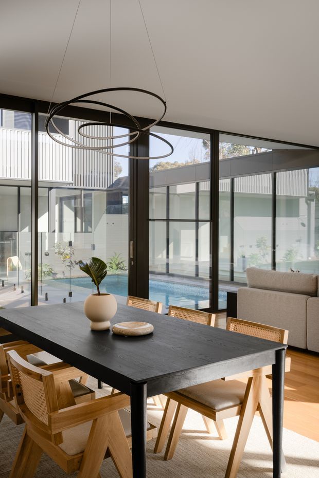 A bright, contemporary dining area with a large dark timber dining table and natural timber chairs, opening to a light-filled living space. Full-height black-framed glazing overlooks an internal courtyard with a swimming pool and modern adjoining architecture. A sculptural circular pendant hangs above the table, while warm timber flooring, a neutral rug, and a beige sofa create a refined, inviting atmosphere.