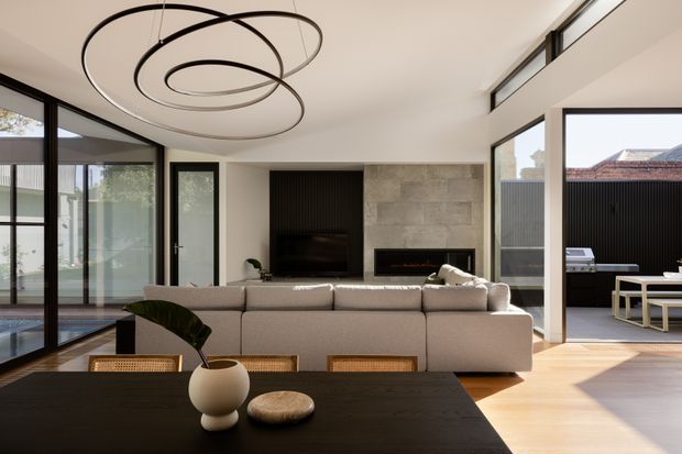 A bright, contemporary open-plan living and dining area with floor-to-ceiling black-framed glazing opening to an outdoor terrace. A large neutral sectional sofa faces a television and linear fireplace set within a dark ribbed feature wall and grey stone surround. The dining table occupies the foreground, while a sculptural looping pendant light, pale timber flooring, and minimalist furnishings create a refined, spacious interior.