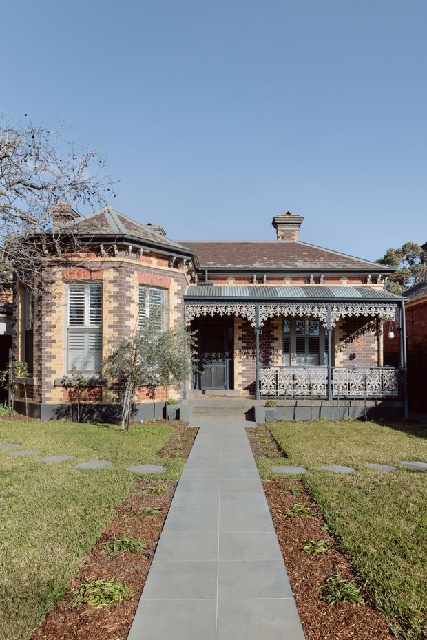 Front exterior view of a single-storey historic brick house with ornate decorative verandah, patterned brickwork, timber shutters, a pitched tiled roof and brick chimney. A straight paved path leads through the front lawn to the raised entry porch.