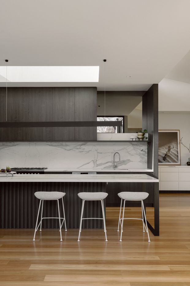 A refined open-plan kitchen dominated by a dark timber kitchen island with vertical fluted detailing, a white veined stone benchtop and matching splashback, and three white bar stools. Warm timber flooring, dark cabinetry, a large skylight, black-framed internal glazing, and understated decorative styling create a contemporary, high-contrast interior.