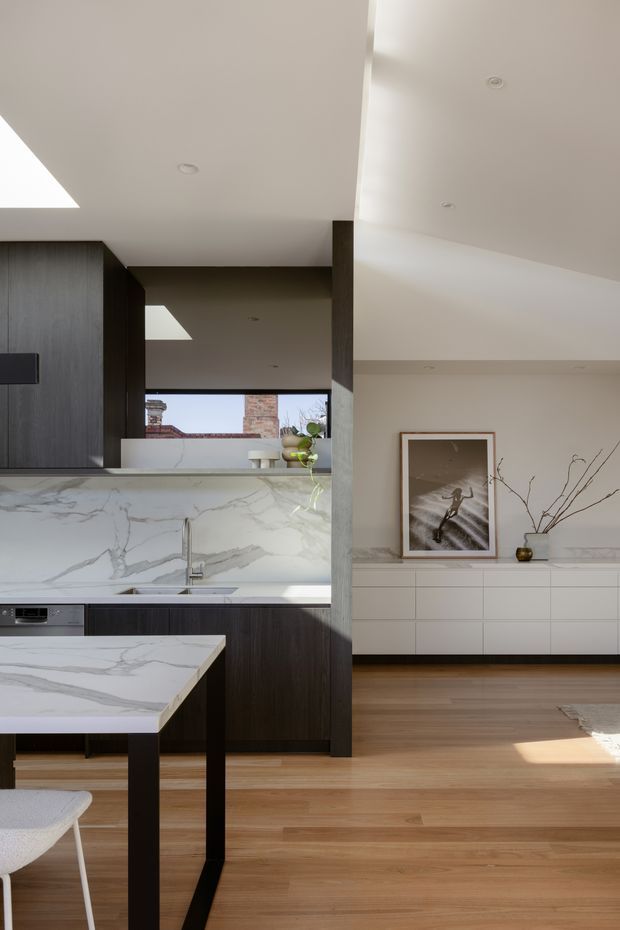 A bright open-plan interior showing a contemporary kitchen and adjacent dining/living area. The kitchen features dark timber cabinetry, a large white marble-look island and splashback, integrated appliances, and a slim undermount sink. Warm timber flooring continues into the adjoining space, which has a long white built-in sideboard, framed artwork, and simple decorative branches. A skylight, recessed ceiling lights, and generous glazing bring in natural light.