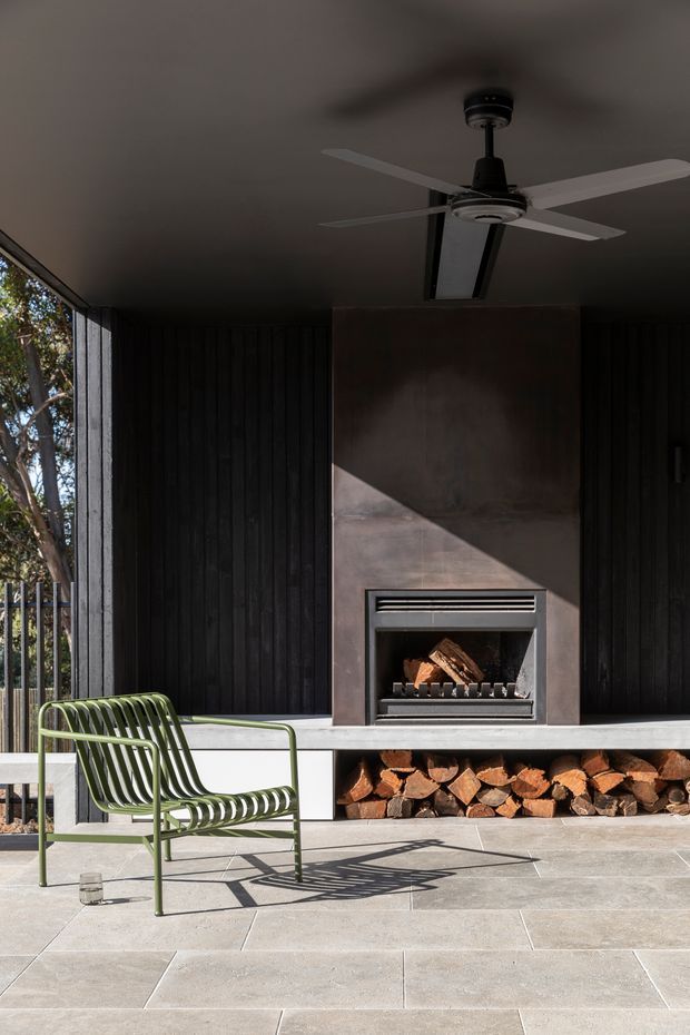 A dark, semi-enclosed outdoor entertaining area with a built-in fireplace and open firewood storage beneath a long concrete bench. Black vertical timber wall cladding, a ceiling fan, pale large-format floor tiles, and a green metal lounge chair define the space. Strong sunlight creates dramatic shadows across the fireplace wall and paving, while trees are visible through the open side.