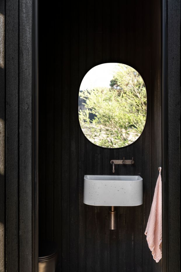 A compact powder room with a wall-mounted oval concrete basin, exposed bronze-toned plumbing, and a round-edged oval mirror. The walls are clad in vertically grooved dark timber, creating a dramatic contrast with the pale basin. A small wall-mounted tap and pink hand towel complete the space, with greenery visible in the mirror reflection.