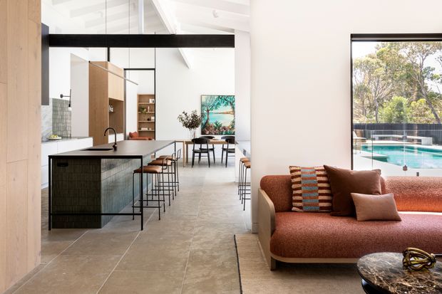 Open-plan contemporary interior combining a kitchen, dining area and living room. The kitchen features a large island with dark textured cabinetry, a pale benchtop and black tapware, while the living area has a rust-coloured sofa and warm timber elements. Full-height glazing connects the space to an outdoor swimming pool, and a dramatic black horizontal structural beam spans the room.