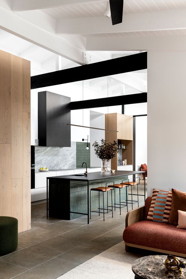 A refined open-plan kitchen and living space with a large dark island, integrated black-framed glazing or mirrors, timber cabinetry, pale stone backsplash, and black exposed ceiling beams. Four bar stools line the island, while a rust-coloured sofa and marble coffee table occupy the foreground. The room has a bright, high-ceilinged feel with strong black, white, timber and warm terracotta contrasts.