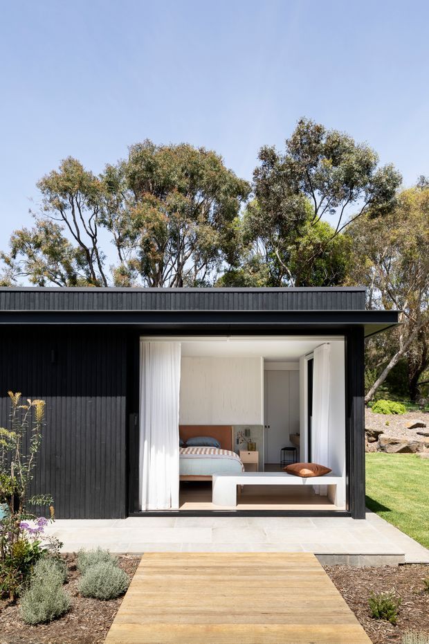 Exterior view of a compact black-clad garden pavilion or dwelling set within a landscaped garden. The single-storey structure has a low flat roof, vertical dark cladding, and a large open glazed opening revealing a simply furnished bedroom with a bed, bedside table, and built-in storage. A timber boardwalk leads to a pale paved threshold, with established trees and planting surrounding the building.