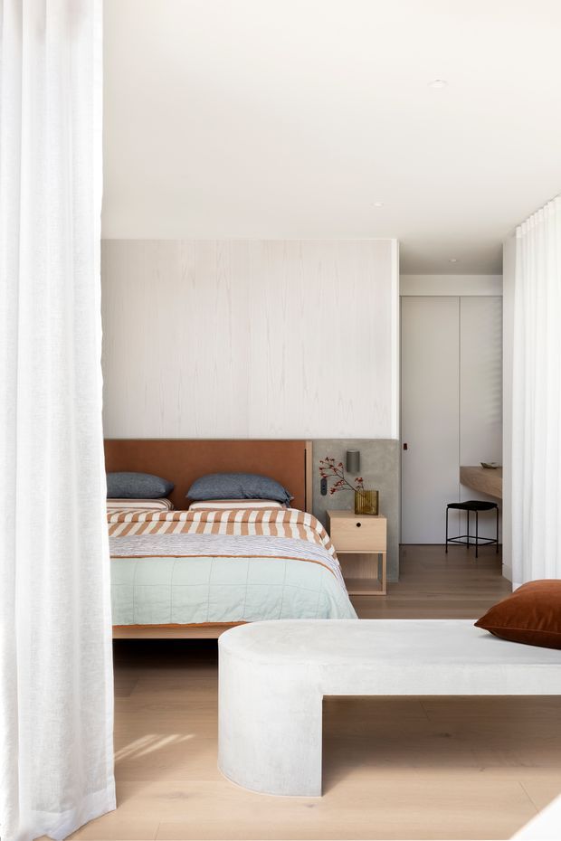 Bright, refined bedroom viewed through sheer white curtains. The room features a low timber bed with a warm brown upholstered headboard, layered blue, striped and pale green bedding, pale timber flooring, white textured wall panelling, integrated wardrobes, a compact timber bedside table, and a large rounded white upholstered bench in the foreground. A small black stool and minimal decor sit near the wardrobe, creating a calm, contemporary atmosphere.