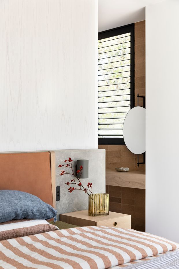 A warmly styled contemporary bedroom featuring a low timber bedside table, a rust-colored upholstered headboard, blue and striped bedding, and a concrete-look bedside wall panel with integrated lighting and power. A large horizontal-louvered black-framed window brings in filtered daylight beside a timber-toned tiled wall and built-in vanity or shelf with a round mirror. Natural timber, concrete, terracotta, and soft neutral tones create a refined, earthy atmosphere.
