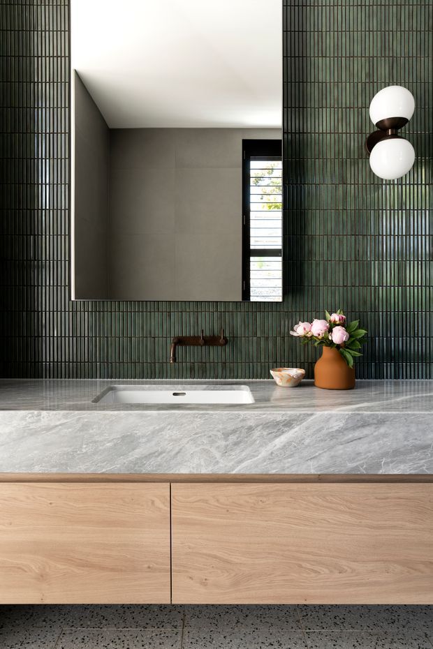 A refined contemporary bathroom vanity featuring a wide grey natural-stone benchtop with an integrated white basin, light timber cabinetry, dark green vertically ribbed wall tiles, a large rectangular mirror, black tapware, and sculptural wall lighting. Terrazzo-style flooring and a vase of flowers add texture and softness.